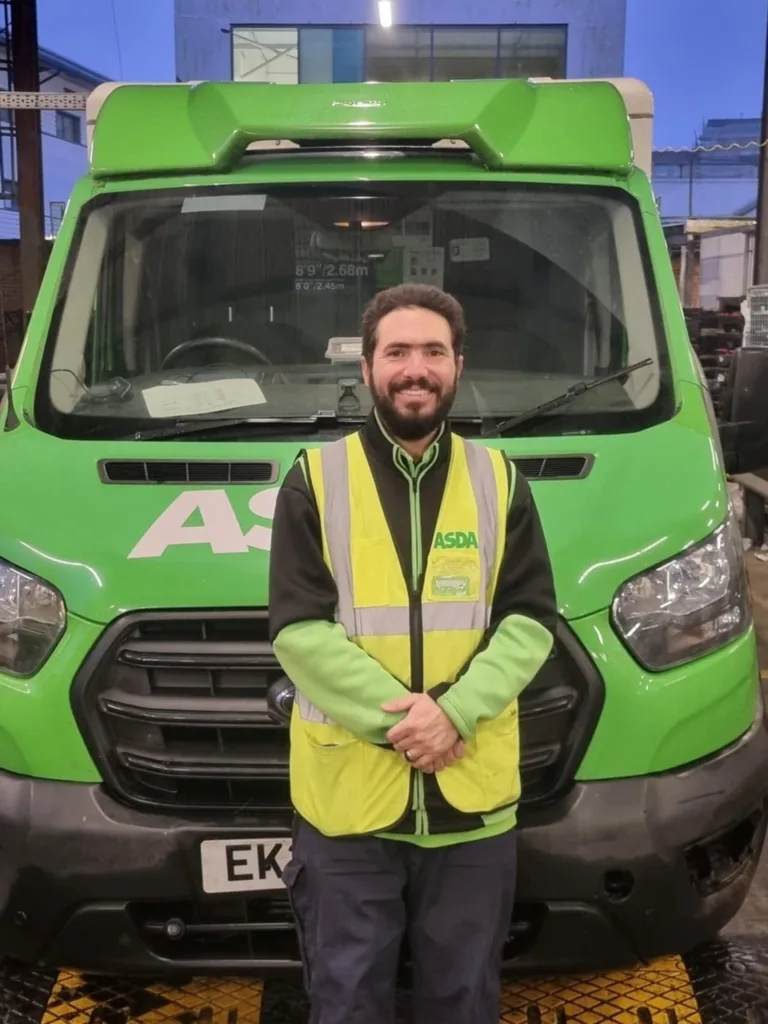 Asda delivery driver Leandro stands smiling in front of his green delivery truck, wearing a high-visibility vest.