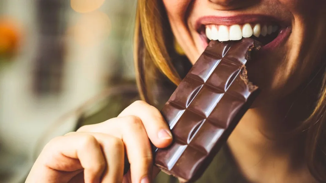 A smiling woman biting into a bar of dark chocolate, enjoying its rich taste.