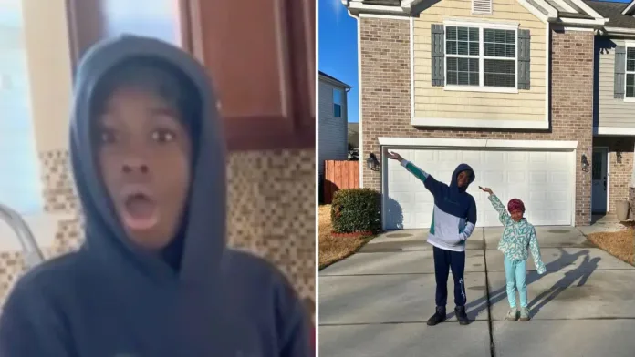 A boy in a hoodie stands in a kitchen with a shocked expression on his face, reacting to the surprise announcement of a new home.