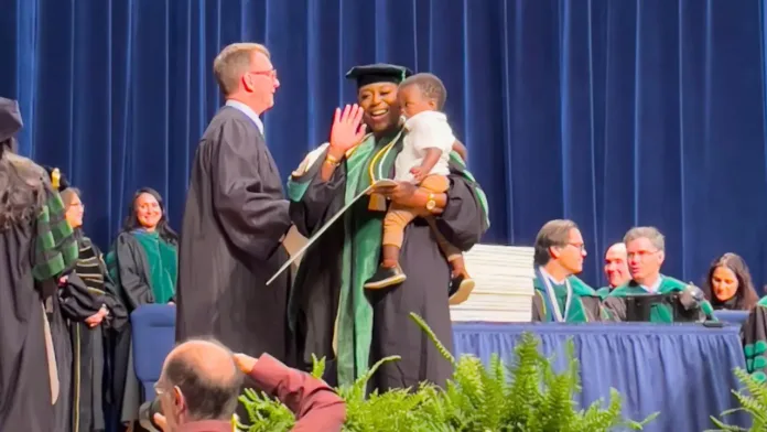 A woman in graduation regalia receives her diploma from a faculty member while holding her toddler son on stage during a commencement ceremony.
