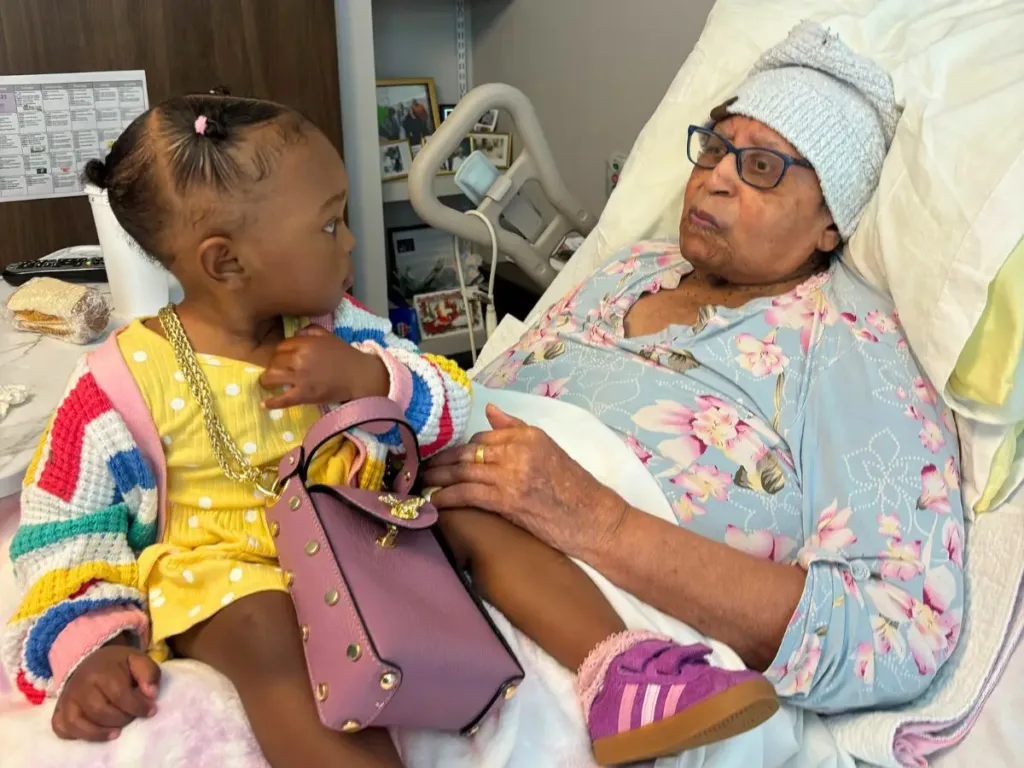 An 18-month-old girl wearing a colorful sweater sits beside her 107-year-old great-great-grandmother, who is resting in bed, as they look at each other thoughtfully.