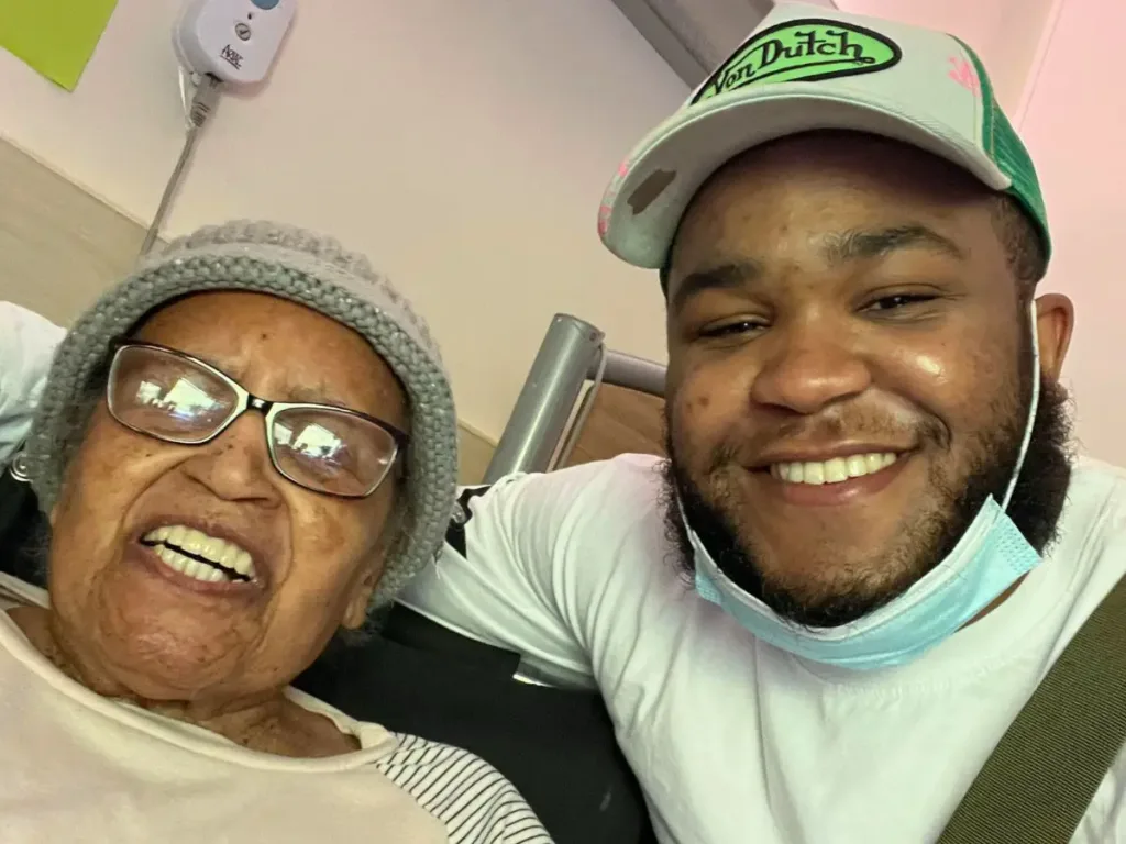 A smiling young man takes a cheerful selfie next to his elderly great-grandmother, who smiles warmly into the camera.