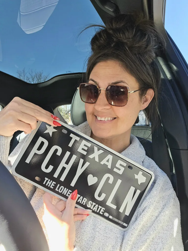 Skye Phillips smiling and holding a customized Texas license plate reading "PCHY CLN" inside a vehicle.