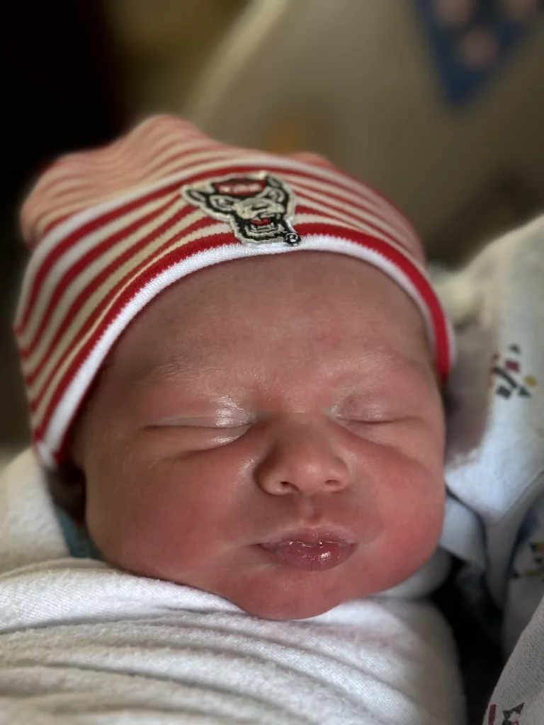 Close-up of a newborn baby wearing a red and white striped hat with the NC State logo, peacefully sleeping wrapped in a hospital blanket.
