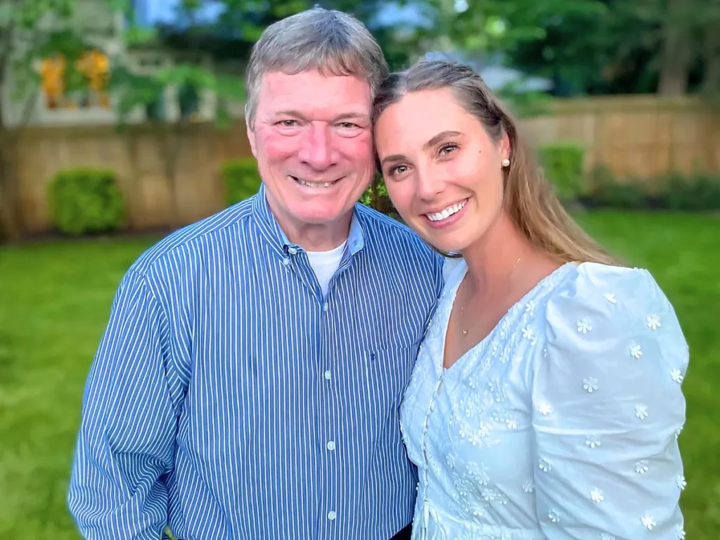 A smiling father and daughter pose together outside in a backyard, standing close and looking happy.