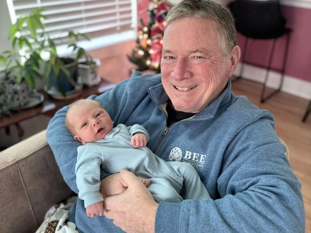 A smiling grandfather holds his newborn grandson while sitting in a warmly lit living room, with holiday decorations in the background.