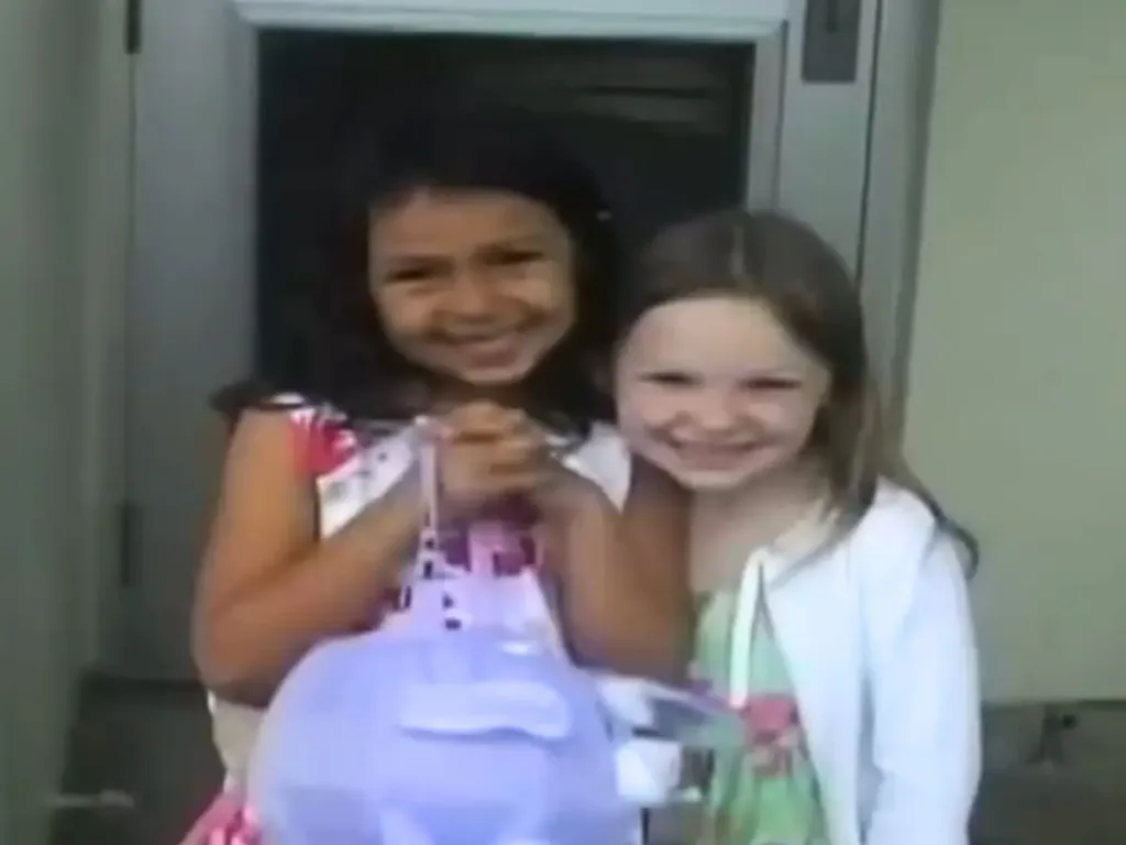Two young girls smiling warmly and embracing each other, standing closely together and appearing very happy.