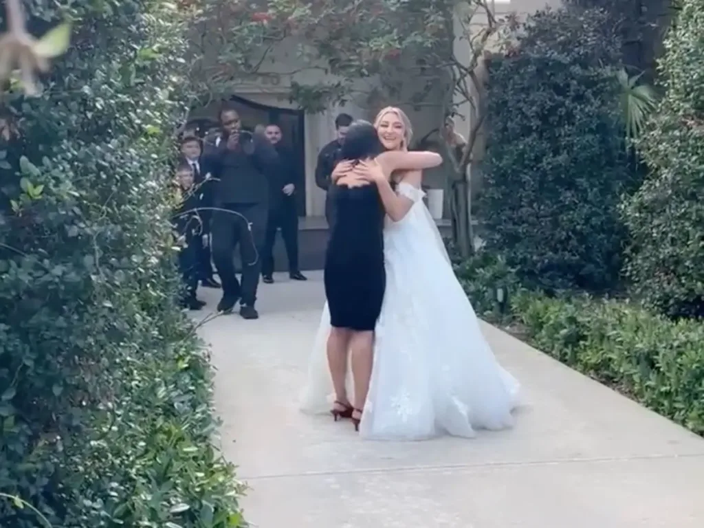 A bride emotionally hugging a friend dressed in black, both women clearly joyful and touched, with wedding guests visible in the background capturing the heartfelt reunion.