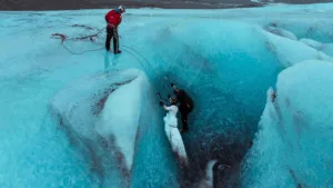 A wide aerial view of a bride and groom in wedding attire climbing a blue glacier ice wall with crampons and ice axes, assisted by a climbing guide standing at the top.
