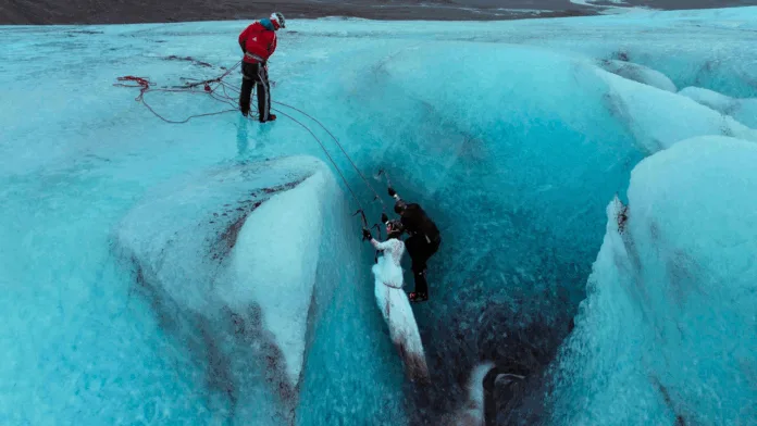 A wide aerial view of a bride and groom in wedding attire climbing a blue glacier ice wall with crampons and ice axes, assisted by a climbing guide standing at the top.