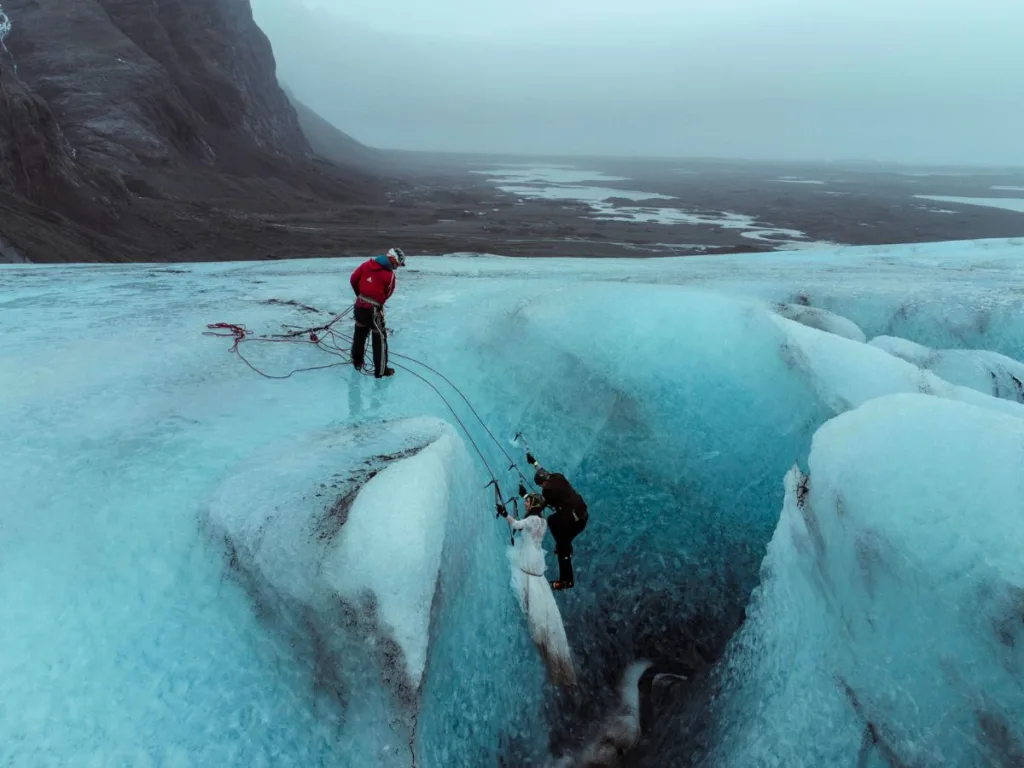 A wide aerial view of a bride and groom in wedding attire climbing a blue glacier ice wall with crampons and ice axes, assisted by a climbing guide standing at the top.