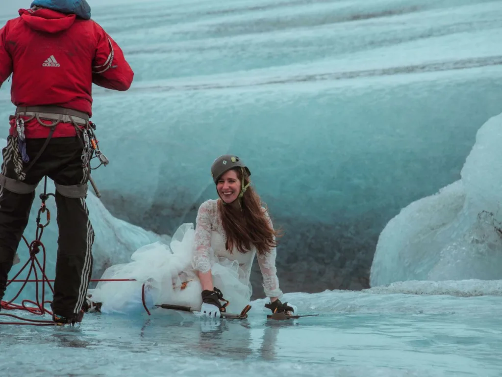 Zoomed-out image showing a dramatic Icelandic landscape with a bride and groom ice climbing out of a deep blue crevasse, assisted by a guide atop the glacier.