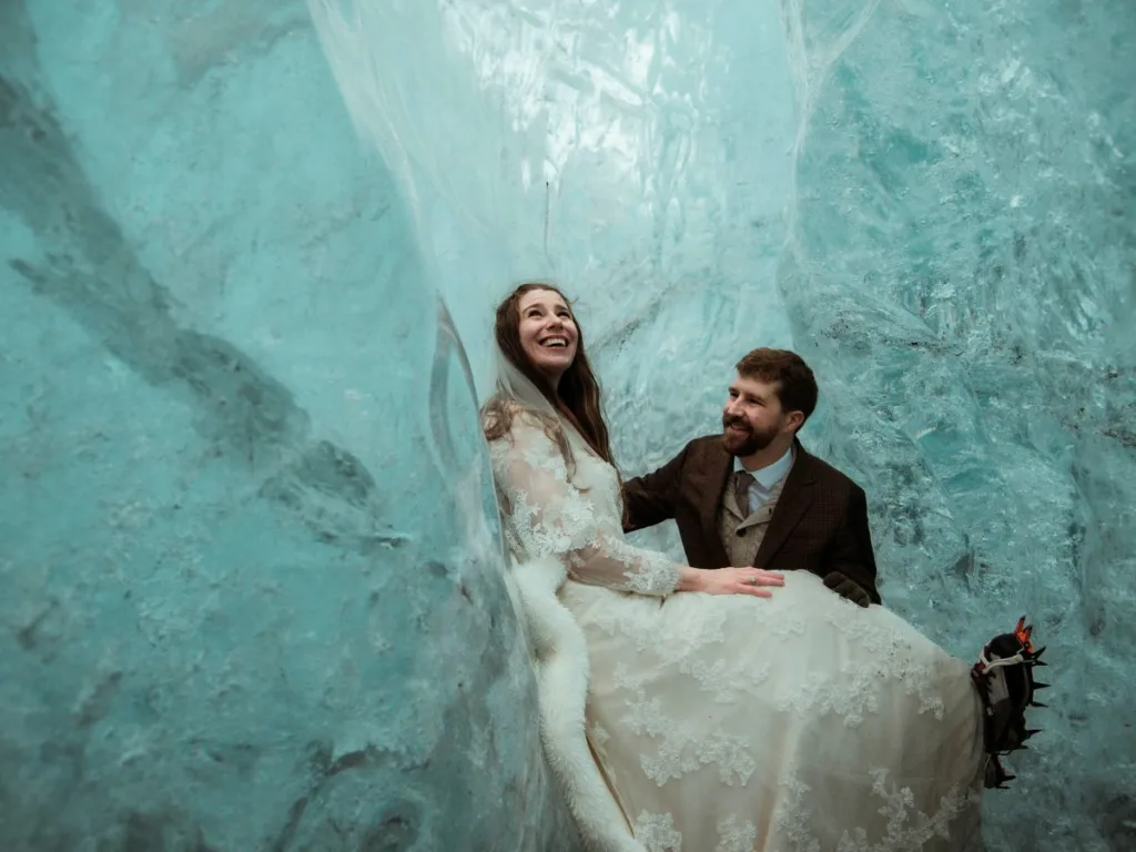 Bride and groom smiling lovingly at each other inside a shimmering blue ice cave, with the bride sitting comfortably on the groom’s lap, still wearing their wedding attire
