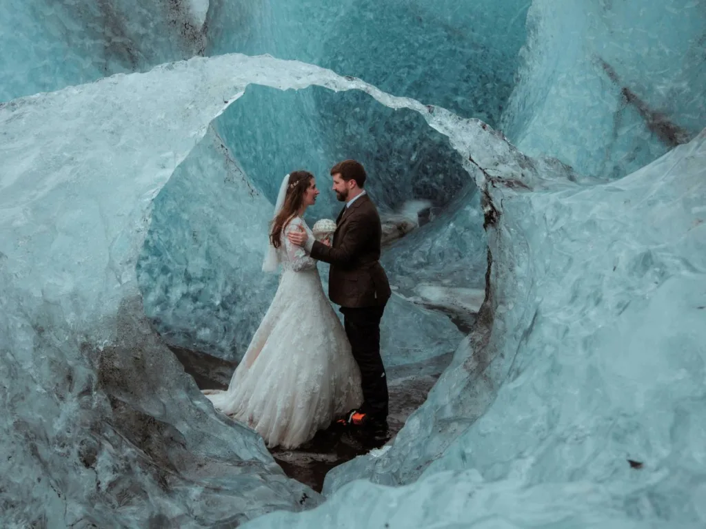 Bride and groom smiling lovingly at each other inside a shimmering blue ice cave, with the bride sitting comfortably on the groom’s lap, still wearing their wedding attire