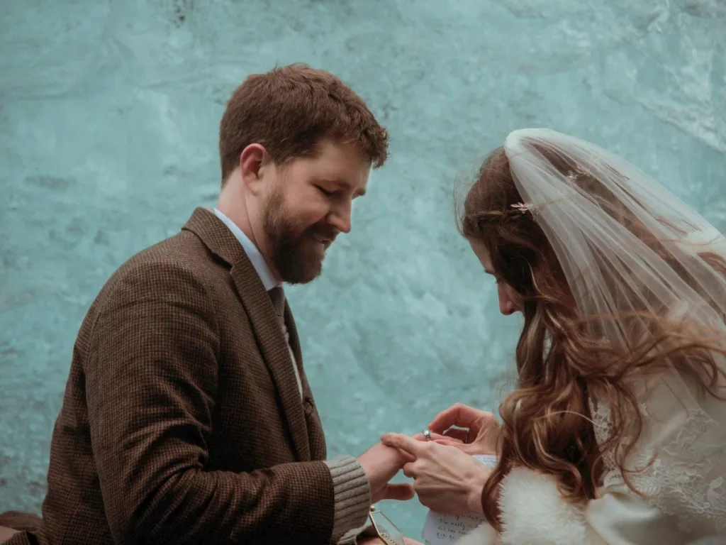 Close-up of the bride placing a wedding ring on the groom’s finger with a soft smile, standing against the textured backdrop of a glacier ice wall.