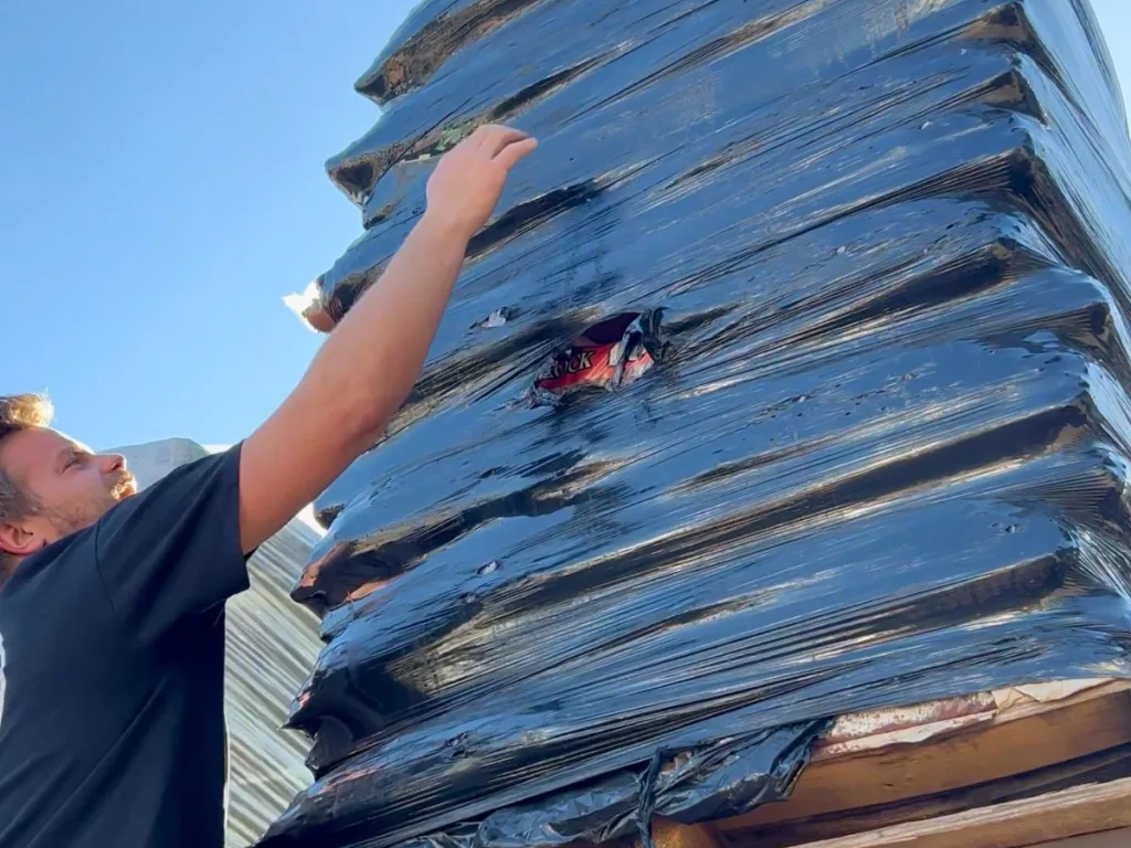 A man stretches his arm toward a shrink-wrapped stack of lava rock bags, inspecting a small opening in the plastic wrap.