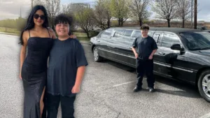 A smiling boy stands proudly next to his sister, who is dressed in a black gown, with a sleek black limousine behind them.