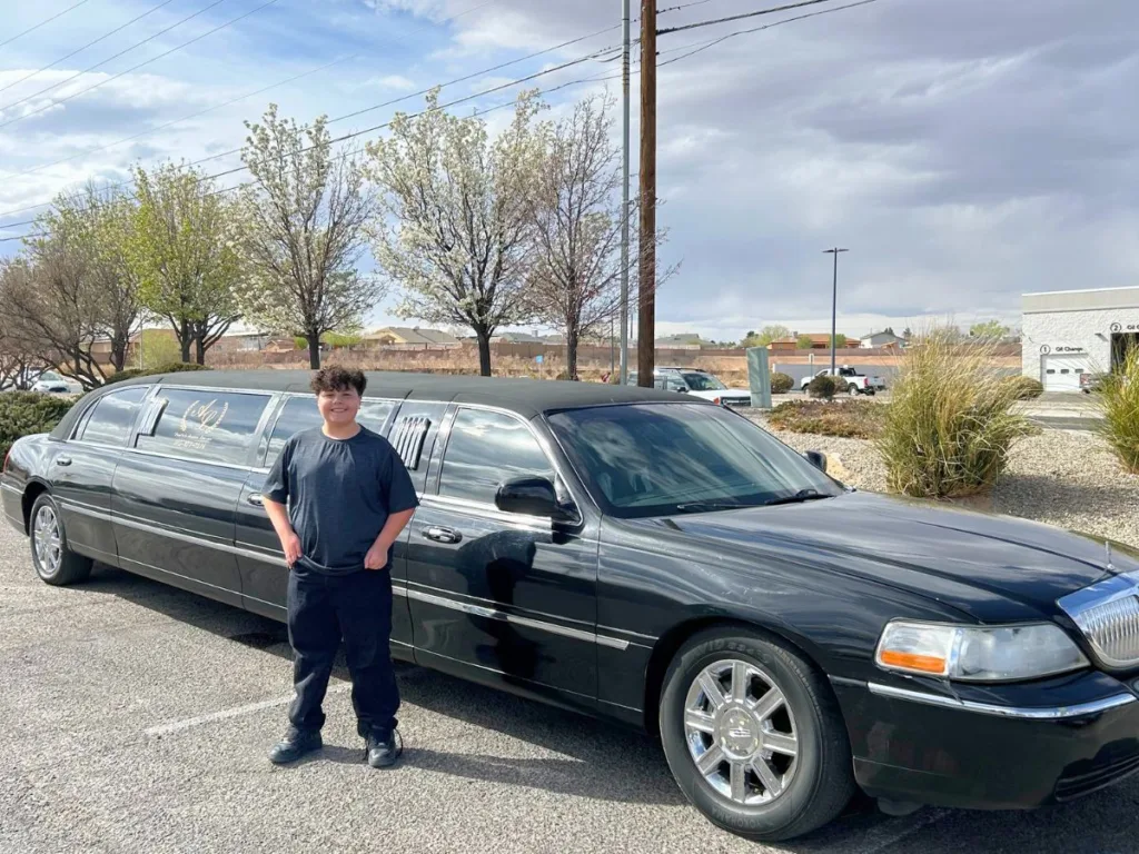 A 12-year-old boy smiles in front of a long black limousine parked under blooming trees in a school parking lot.