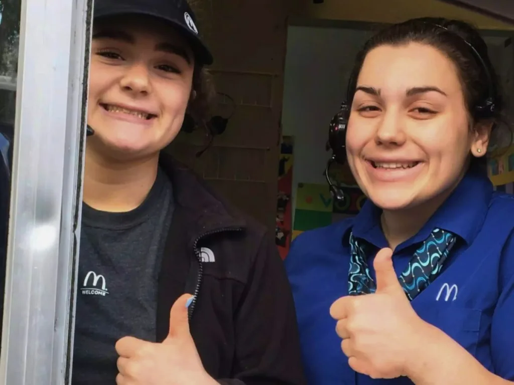 Two smiling McDonald’s employees giving thumbs-up at a drive-thru window.
