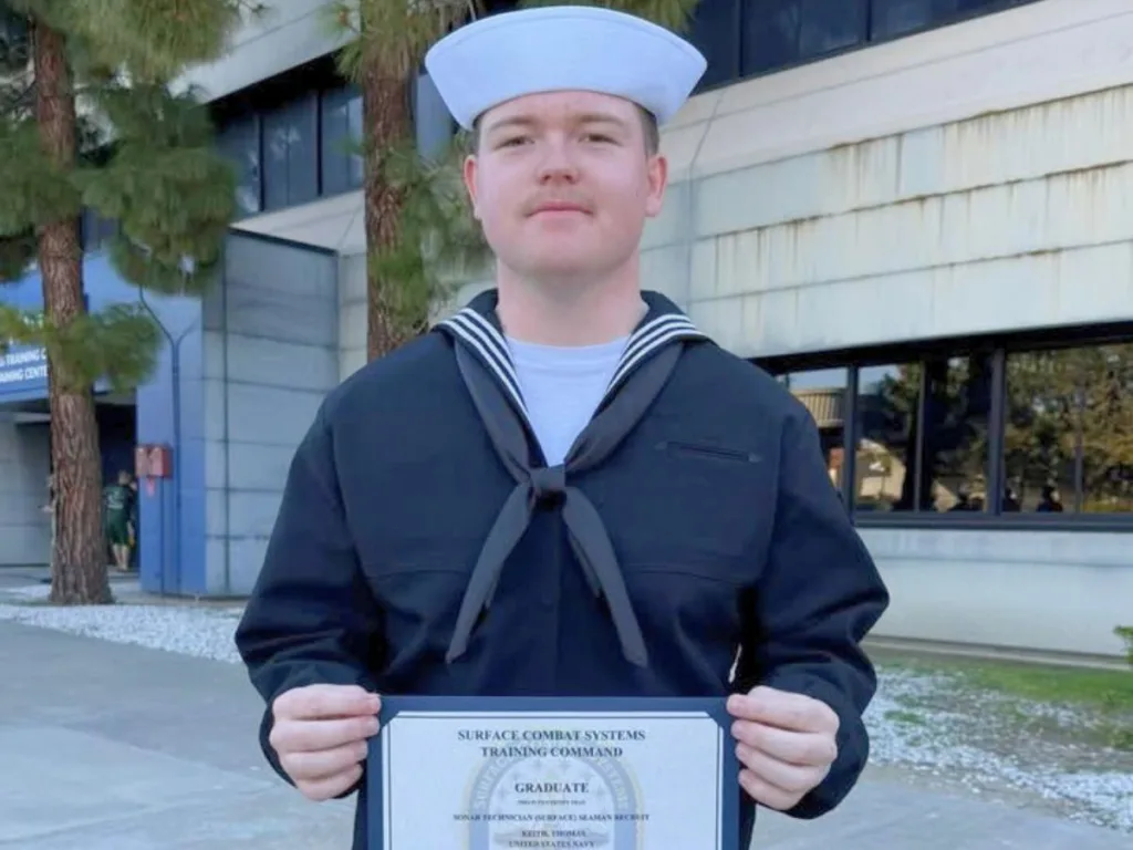Navy graduate in uniform holding his Surface Combat Systems Training Command certificate.