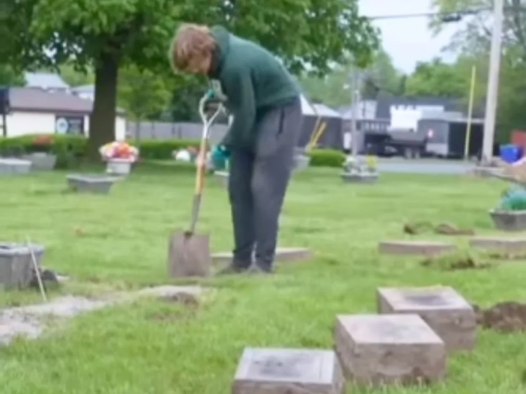 A high school student using a shovel to dig and raise a sunken grave marker at Mount Calvary Cemetery.