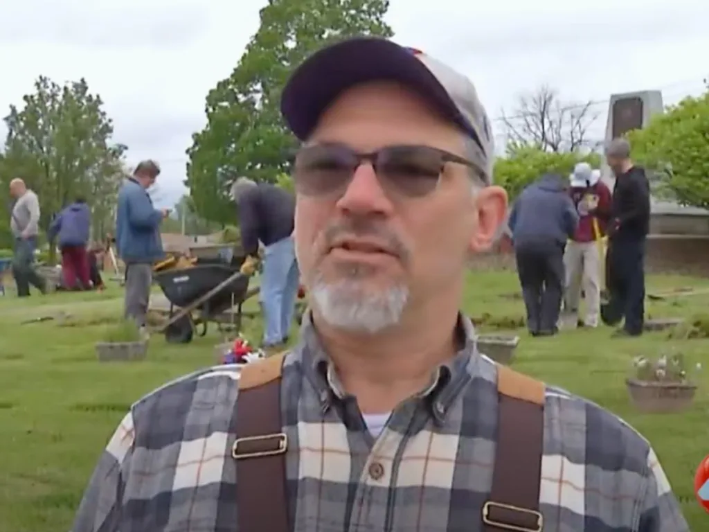 Paul Mueller, the project organizer, wearing a cap and glasses, speaks while volunteers work in the background at the cemetery.