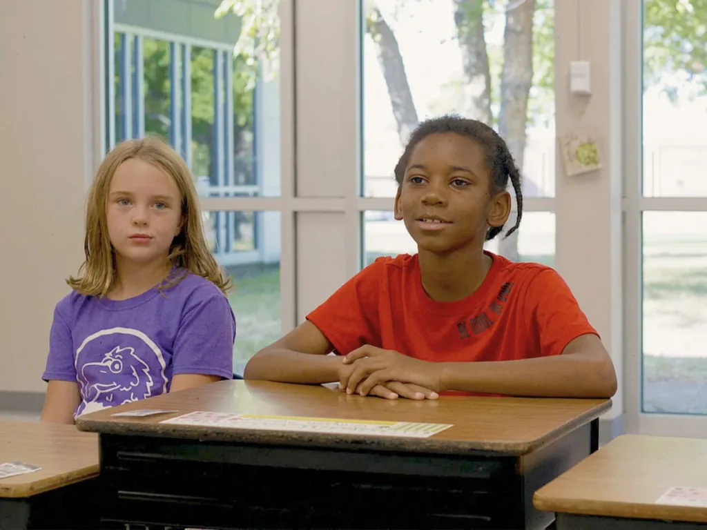 Two young students sitting at their desks in front of large windows letting in daylight.