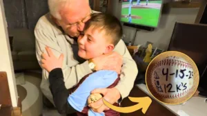 Young boy hugs elderly man while holding a baseball, with an inset showing the ball labeled “4-15-25, 12 K’s.”