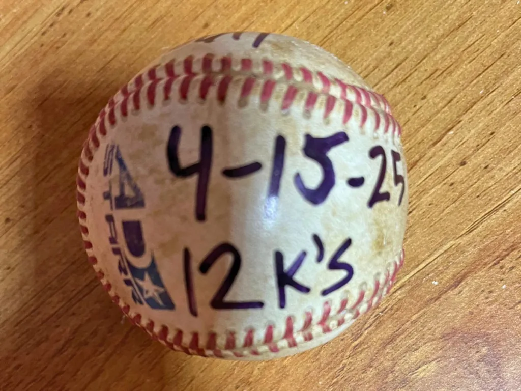 Close-up of a baseball marked “4-15-25, 12 K’s” on a wooden table.