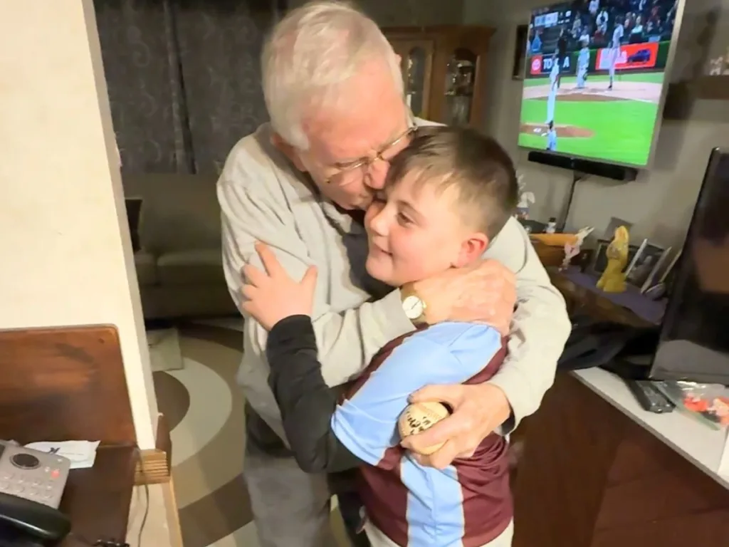 Elderly man embraces young boy holding a baseball inside a living room.