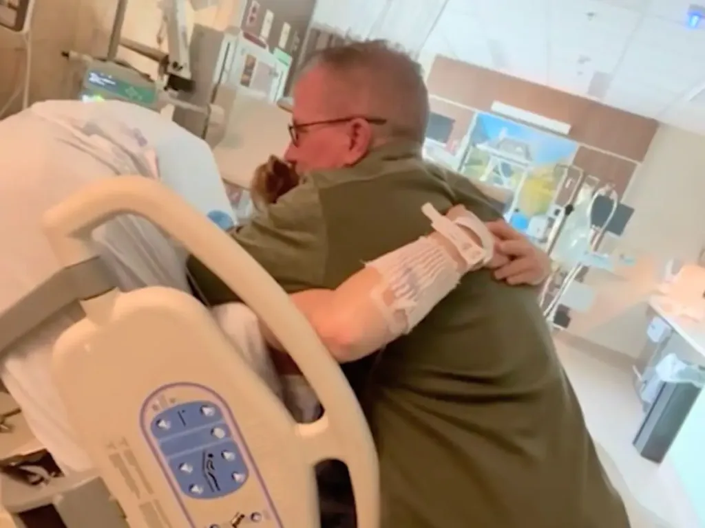 Stepdad leans over a hospital bed to hug his stepdaughter after the baby name reveal.