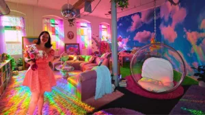 Woman holding flowers and smiling in a vibrant, rainbow-lit loft filled with colorful furniture and decor.