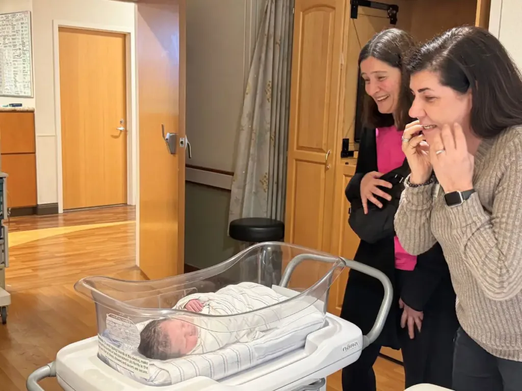 Natalie and Joanna stand side by side in a hospital hallway, mouths open in happy surprise as they glimpse newborn Monroe.