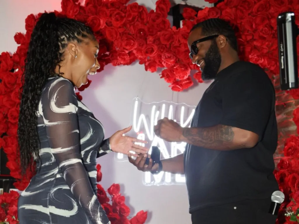 A woman reacts with shock and delight while a man holds an open ring box, red rose decorations in the background.