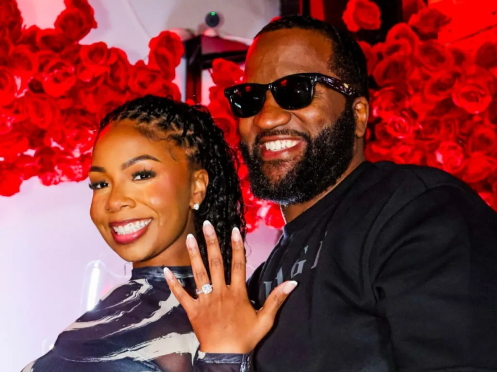 The newly engaged couple smile in front of a wall of red roses, the woman showing off her left hand to display the engagement ring.