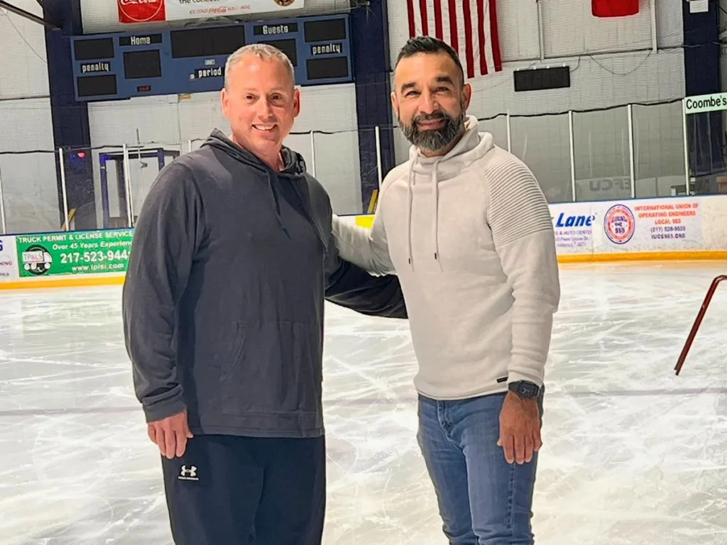 Two smiling men pose on an ice rink after reuniting as Navy friends.