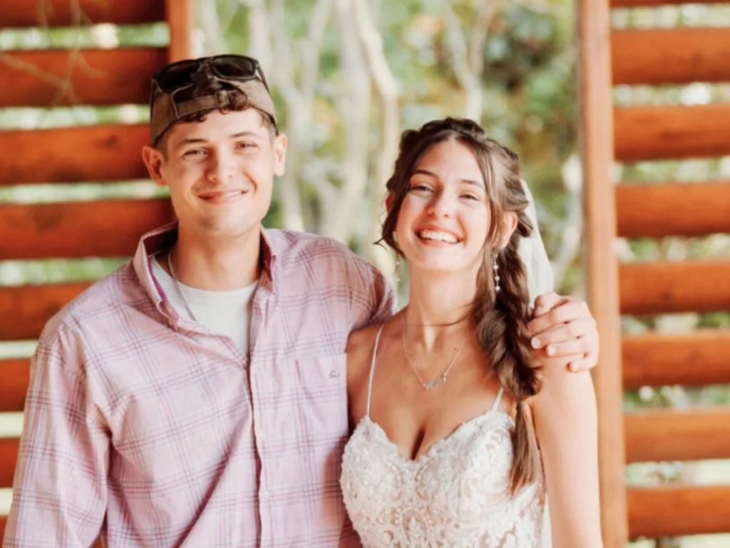 Bride and man in cap smile with arms around each other on wedding day.