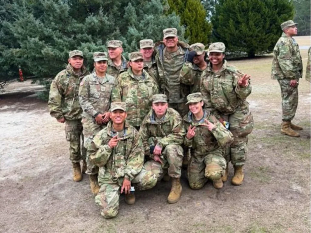 A group of uniformed soldiers kneel and stand together, smiling and flashing peace signs in front of evergreen trees.