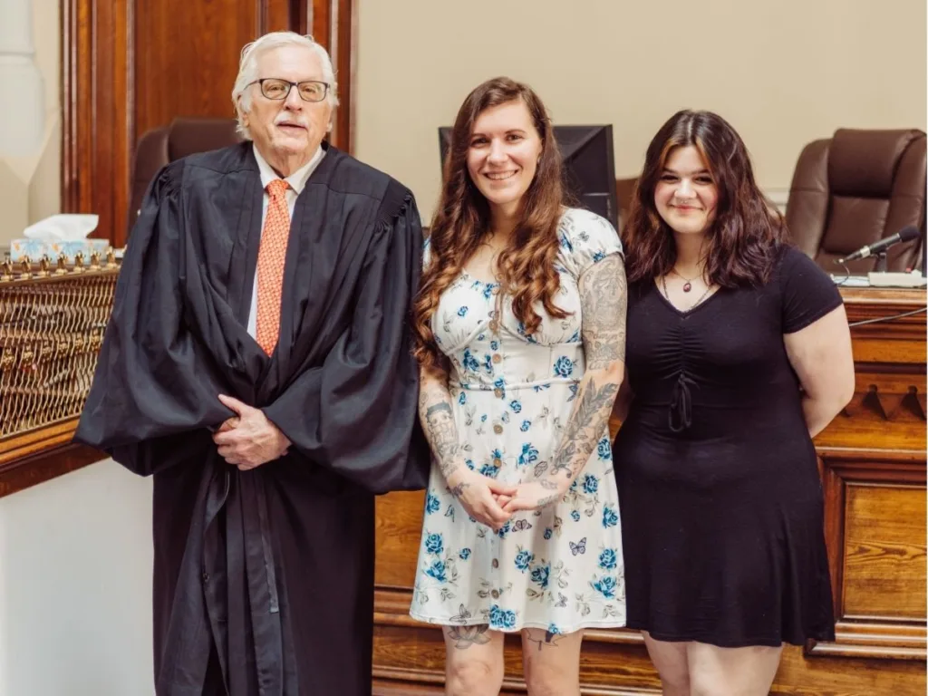 Judge stands with an adoptive mom and her teen daughter smiling inside a courtroom after the adoption hearing.