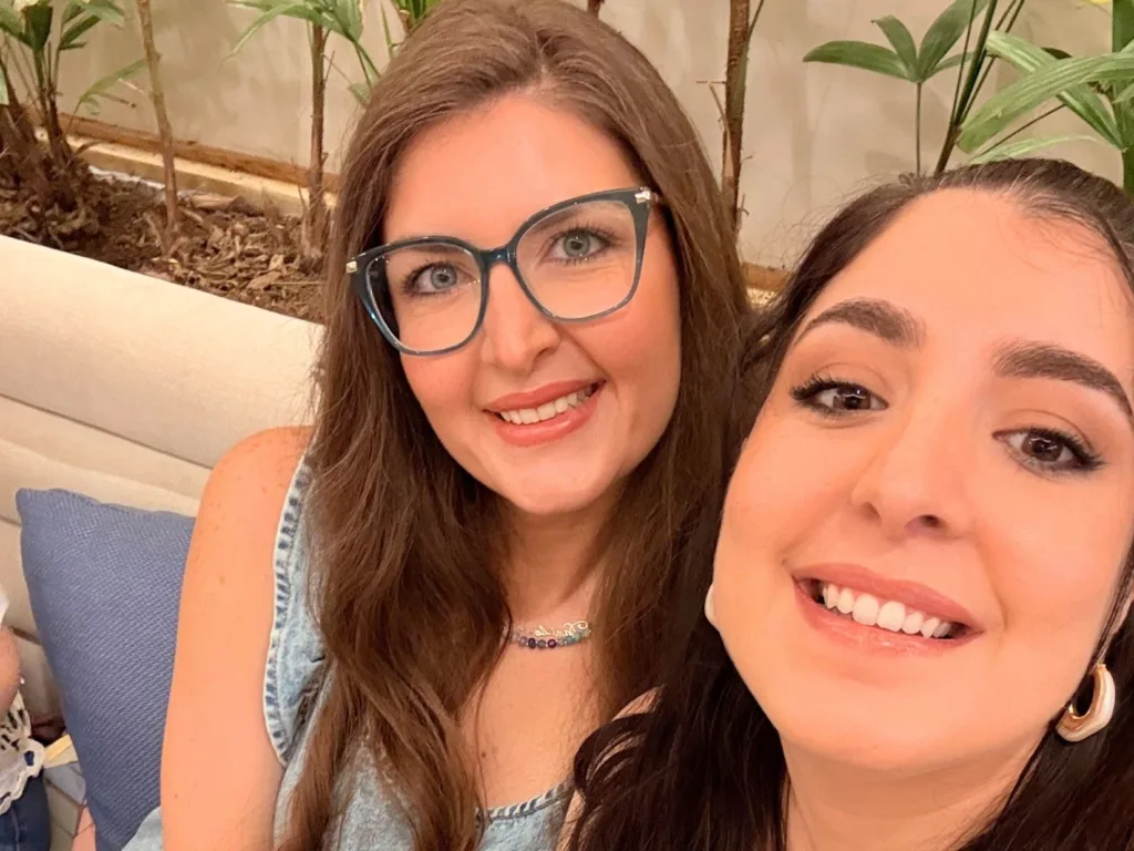 Close-up selfie of two sisters smiling on a sofa, celebrating the pregnancy announcement that echoes their family tradition.