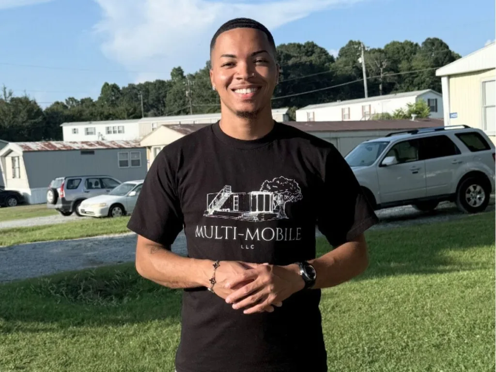 Tyquan Carr smiles in a T-shirt reading “Multi-Mobile LLC” while standing in a mobile home community.