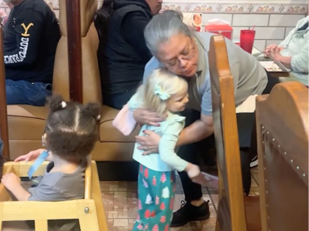 Server with gray hair embracing young blonde child in patterned skirt at restaurant booth