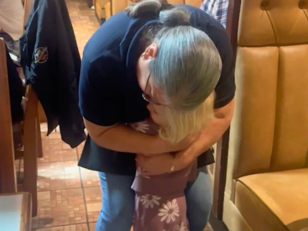 Woman and young girl hugging tightly in restaurant dining area with wooden chairs