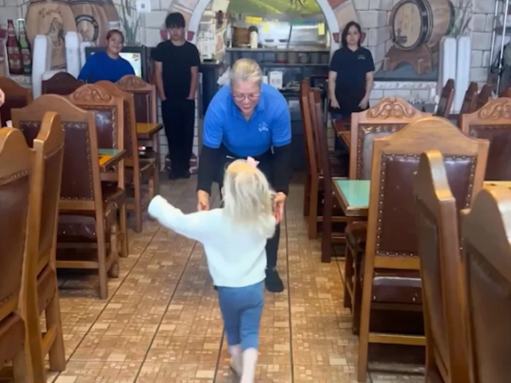 Toddler approaching crouching server in blue shirt with restaurant staff watching in background.