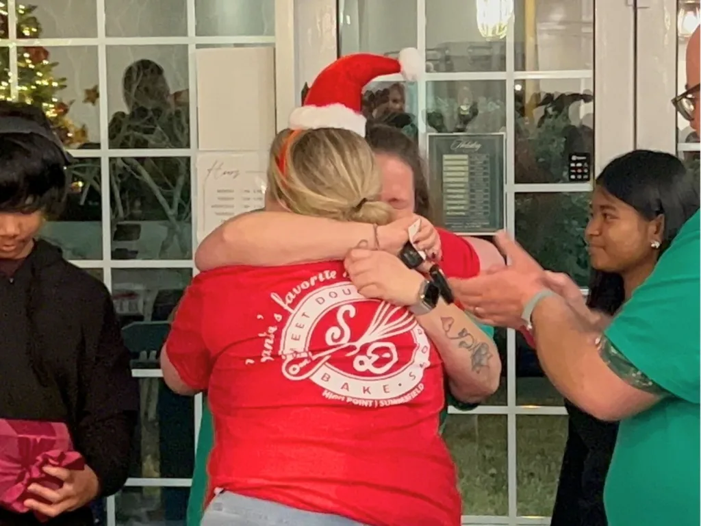 A woman in a red shirt hugs someone tightly as coworkers clap during a holiday party.
