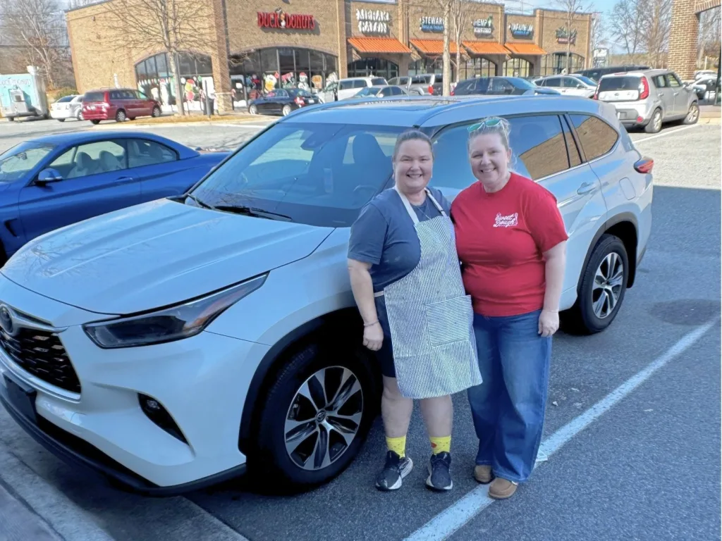 Two women smile while posing beside a white SUV in a parking lot outside a row of shops.