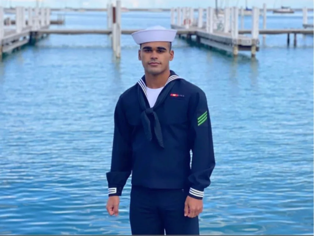 Chris stands in his U.S. Navy uniform near a waterfront dock.