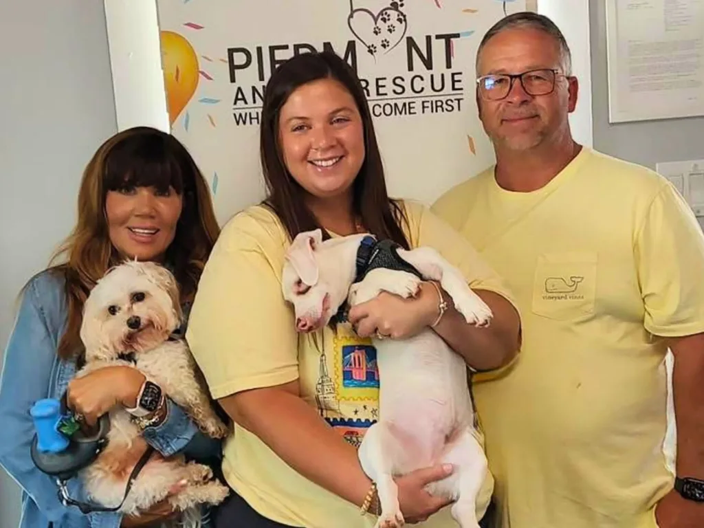 Three people pose inside an animal rescue while one holds a small fluffy white dog and another cradles a white dog wearing a black harness.
