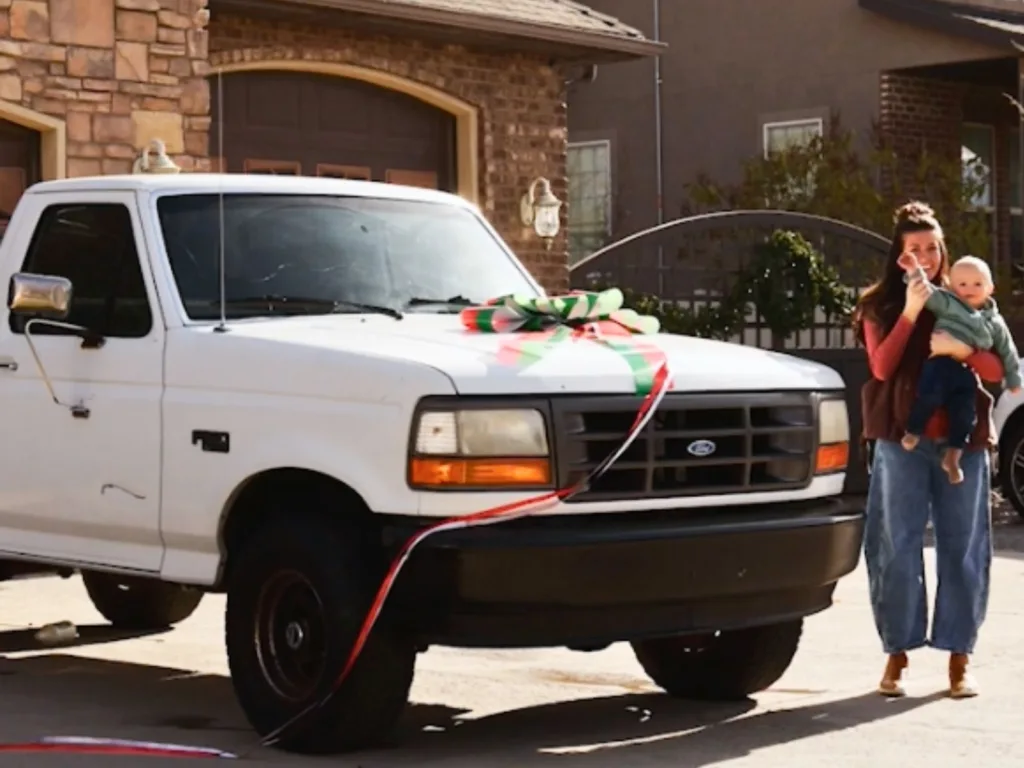 White Ford F-150 with a large gift bow on the hood parked in a driveway as a woman holds a baby.