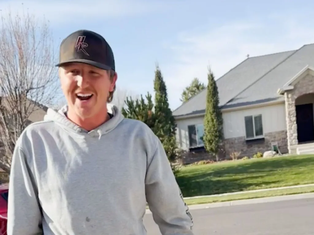 Smiling man in a gray hoodie and black cap standing on a suburban driveway